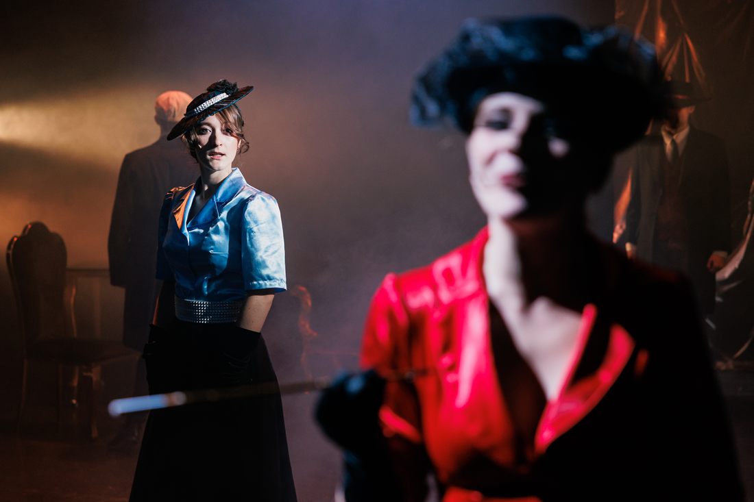Žena v modré halence a klobouku, v rozostřeném popředí žena v červeném